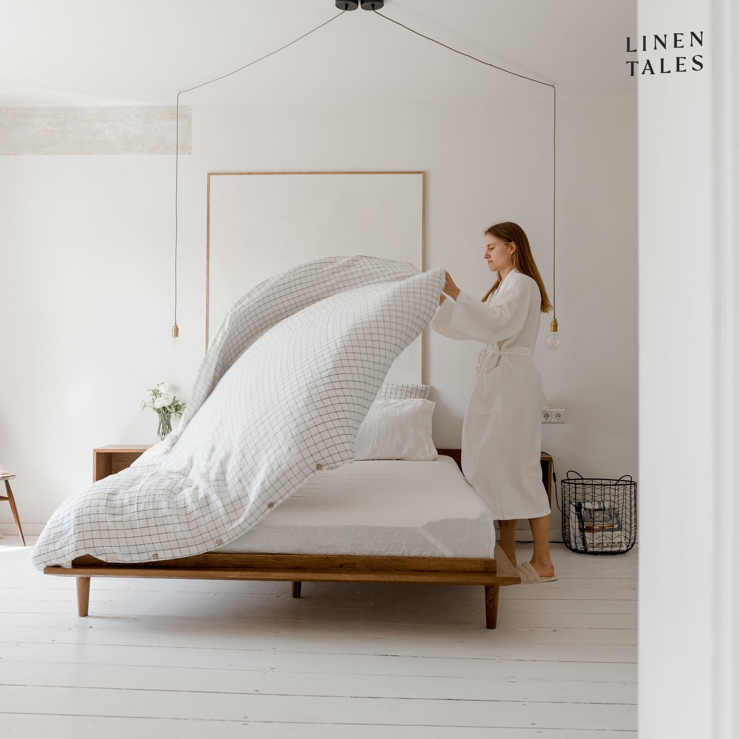 A woman gracefully floats a crisp white grid duvet cover over a fully made linen bed, bringing effortless elegance and comfort.