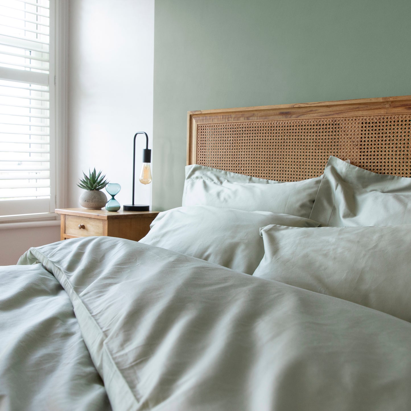 Neatly made bed with light-colored seagrass green bedding, including duvet cover, pillowcase and fitted bed sheets, and a wooden headboard in a bright bedroom.