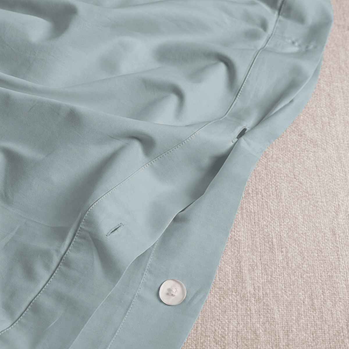 Close-up of a light sage green button-up shirt laid flat on a textured beige fabric surface, showing the fabric texture, stitching, and a white button.