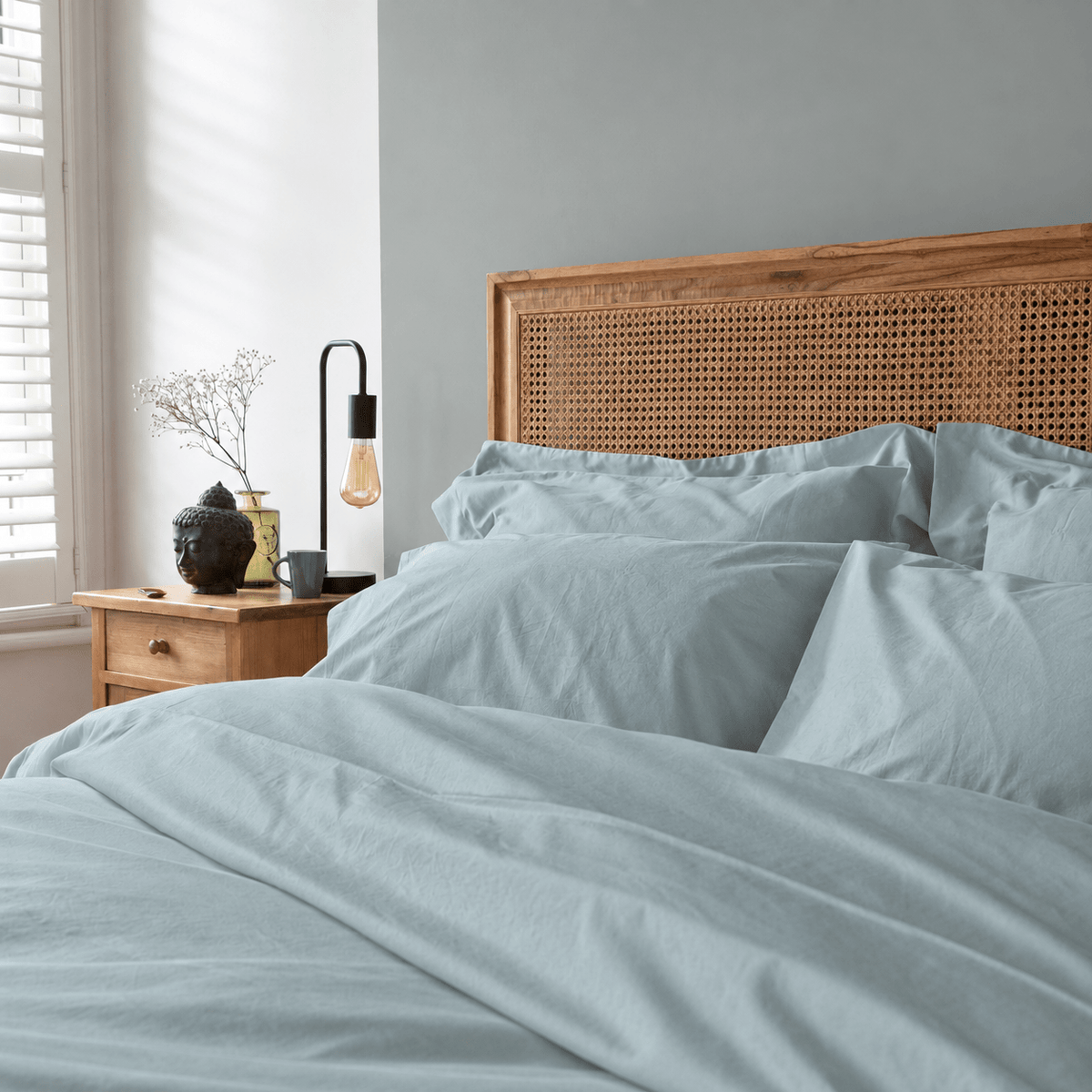 Light blue bedding set with matching pillows and duvet, styled in a bedroom setting with a wooden cane headboard, bedside table, and lamp.