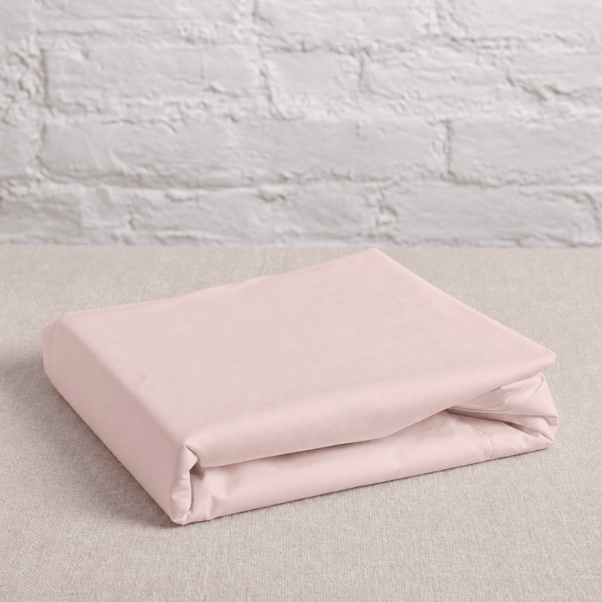 A folded light pink cotton flat sheet on a textured beige surface against a white brick wall background.