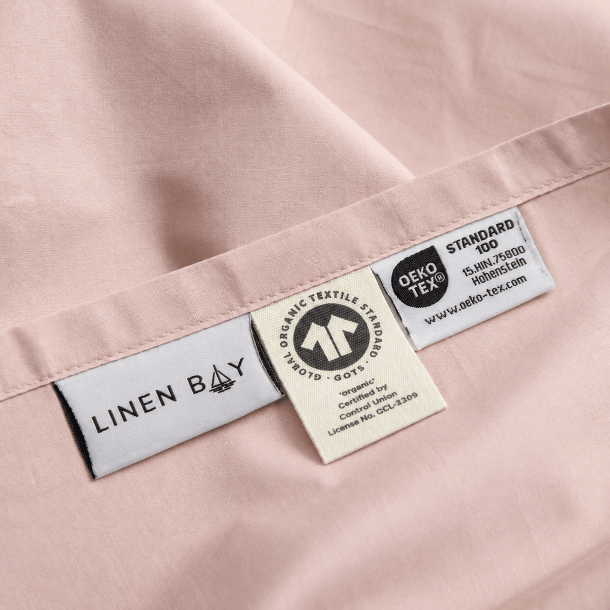 Close-up of a pale blush pink fabric garment showing sewn-in labels, including LINEN BAY and DEKO TEX certification tags.