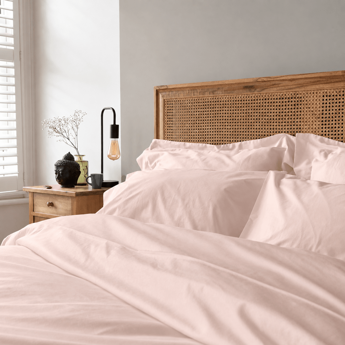 A neatly made bed with pale blush pink bedding and matching pillows, set in a softly lit bedroom with a wooden cane headboard, bedside table, lamp, and window.