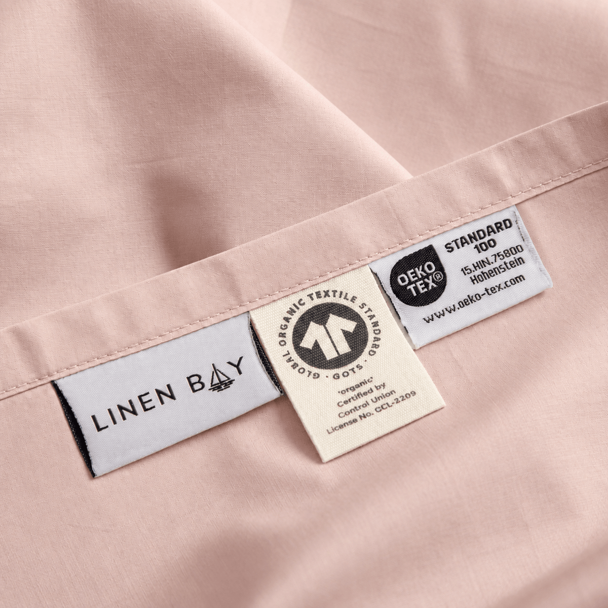 Close-up of a pale blush pink fabric garment showing sewn-in labels, including LINEN BAY, OEKO-TEX Standard 100, and Organic Textile Standards.
