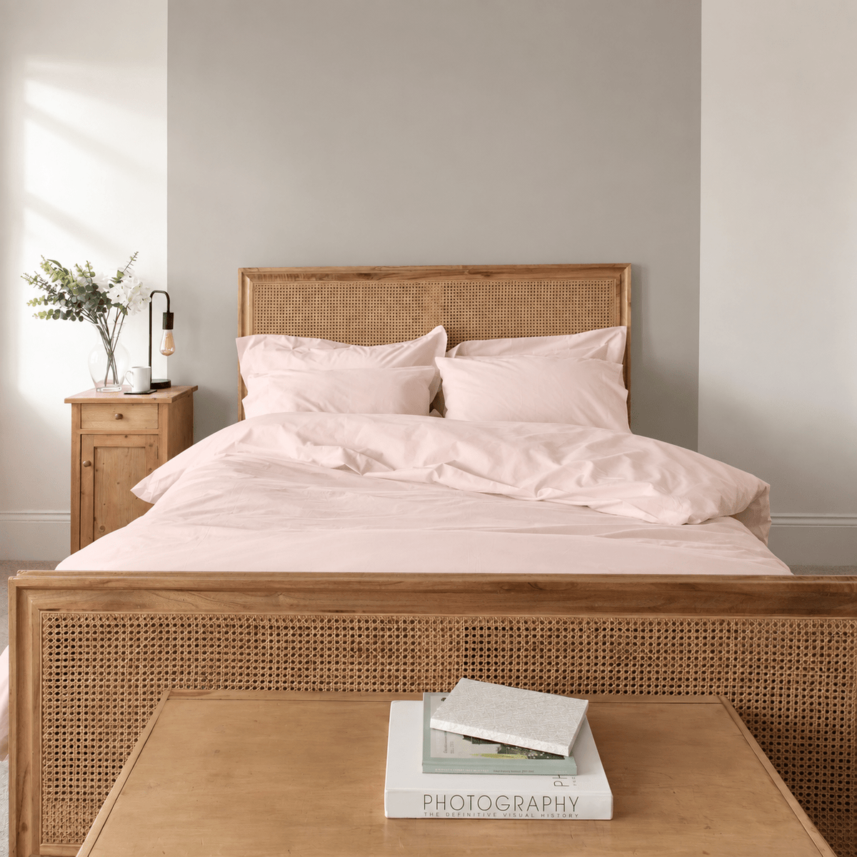 A neatly made bed with pale blush pink bedding and matching pillows, set in a minimalist bedroom with a light wood rattan headboard and footboard, a wooden nightstand with a lamp, and a low wooden table in the foreground holding books.
