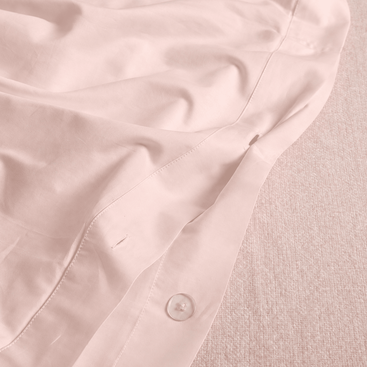 A close-up of a pale blush pink button-up shirt fabric laid over a textured beige fabric background, showing the smooth satin-like material and a single button.