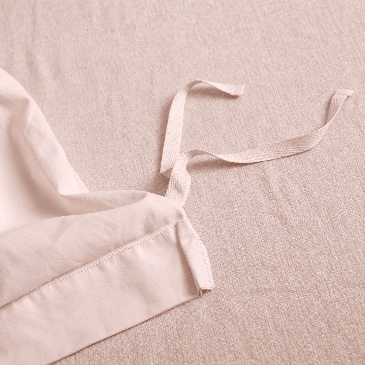 Close-up of a light blush pink fabric garment detail with a tie strap on a matching textured fabric background.