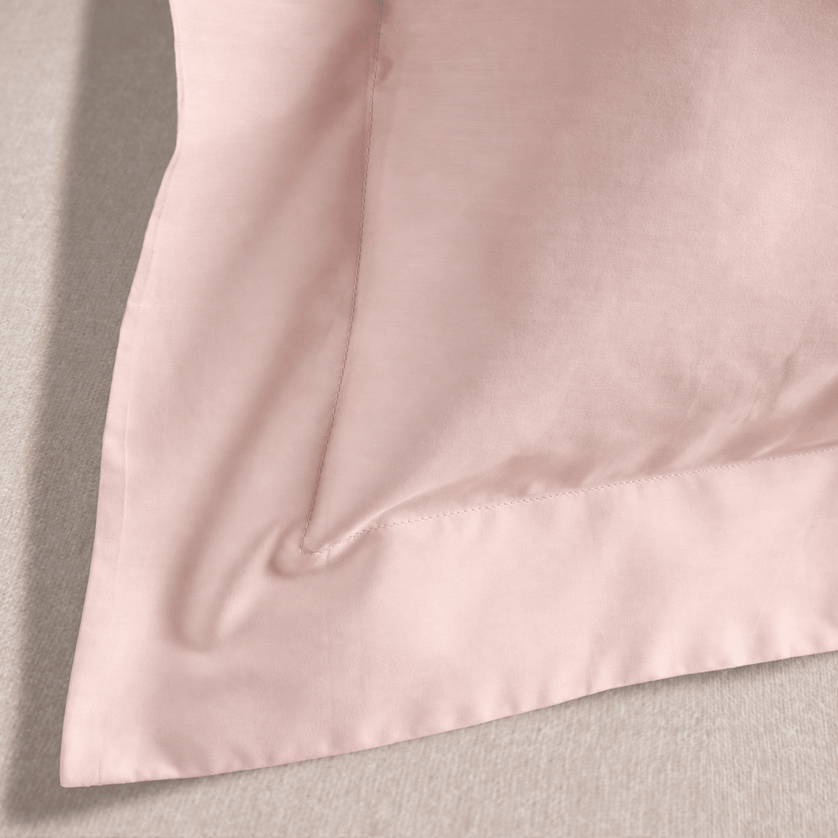 A close-up of a light pink satin or silk pillowcase corner with a stitched border, laid flat on a textured gray surface.