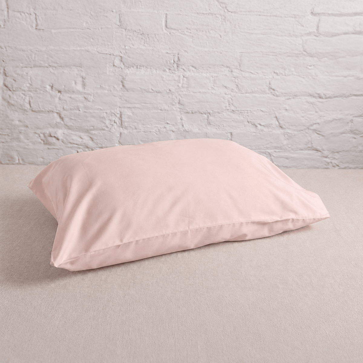 A light pink pillowcase resting on a beige textured surface against a white brick wall background.