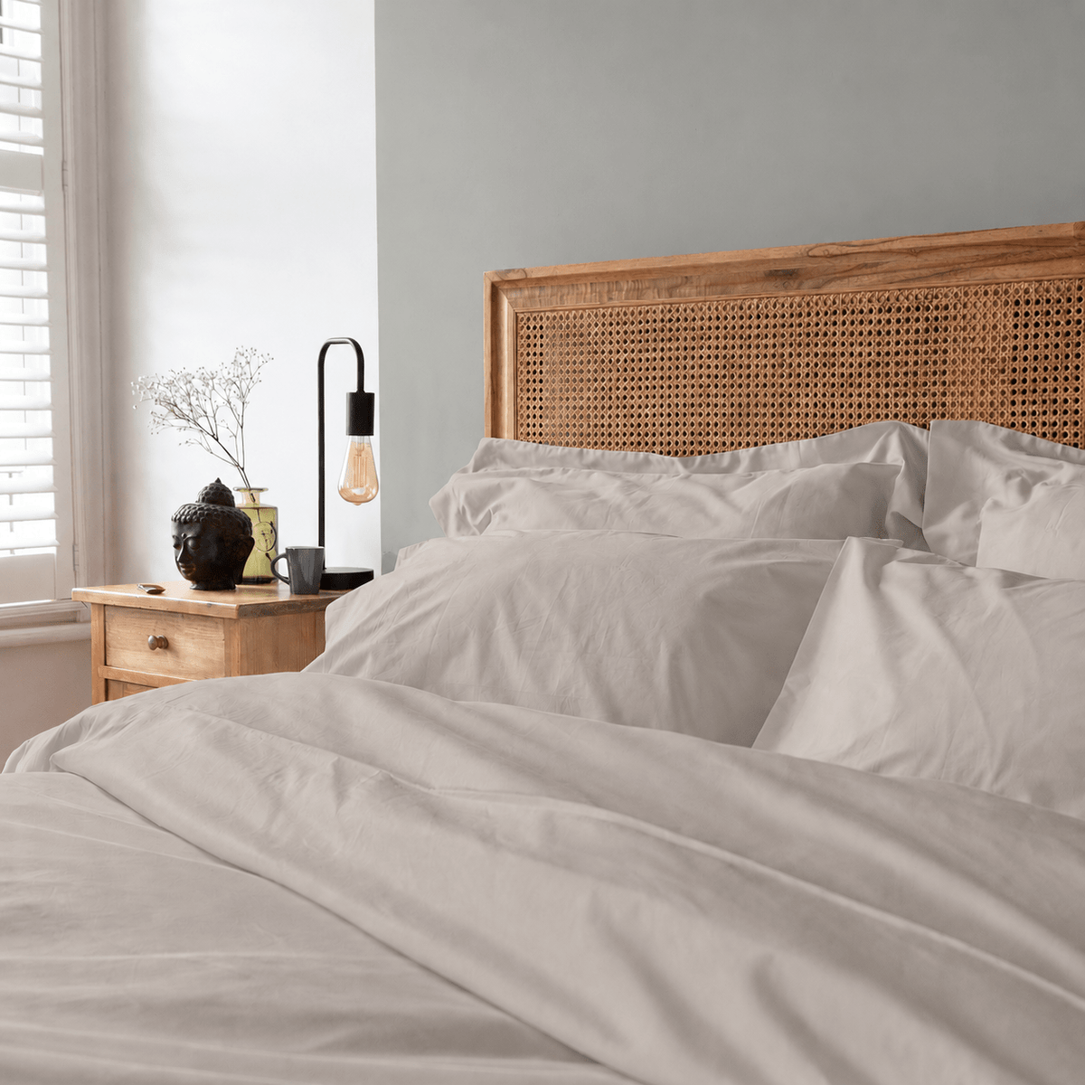 A neatly made bed with harbour stone bedding and a wooden cane headboard in a softly lit bedroom. A bedside table with a black lamp and small vase sits beside a window with white shutters.