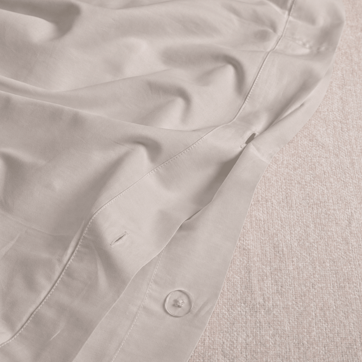 A close-up of a light grey fabric garment, showing the texture of the material, a button, and the folded edges of the fabric against a textured neutral background.