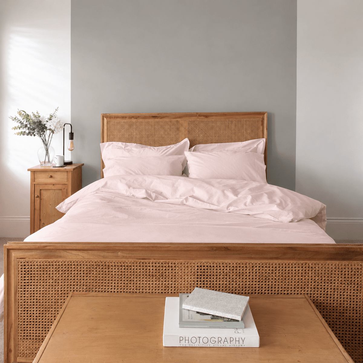 A neatly made bed with light pink bedding and matching pillows, set in a bedroom with a wooden cane headboard and matching footboard. A wooden nightstand with a lamp and flowers is on the left, and a wooden table with books is in the foreground.