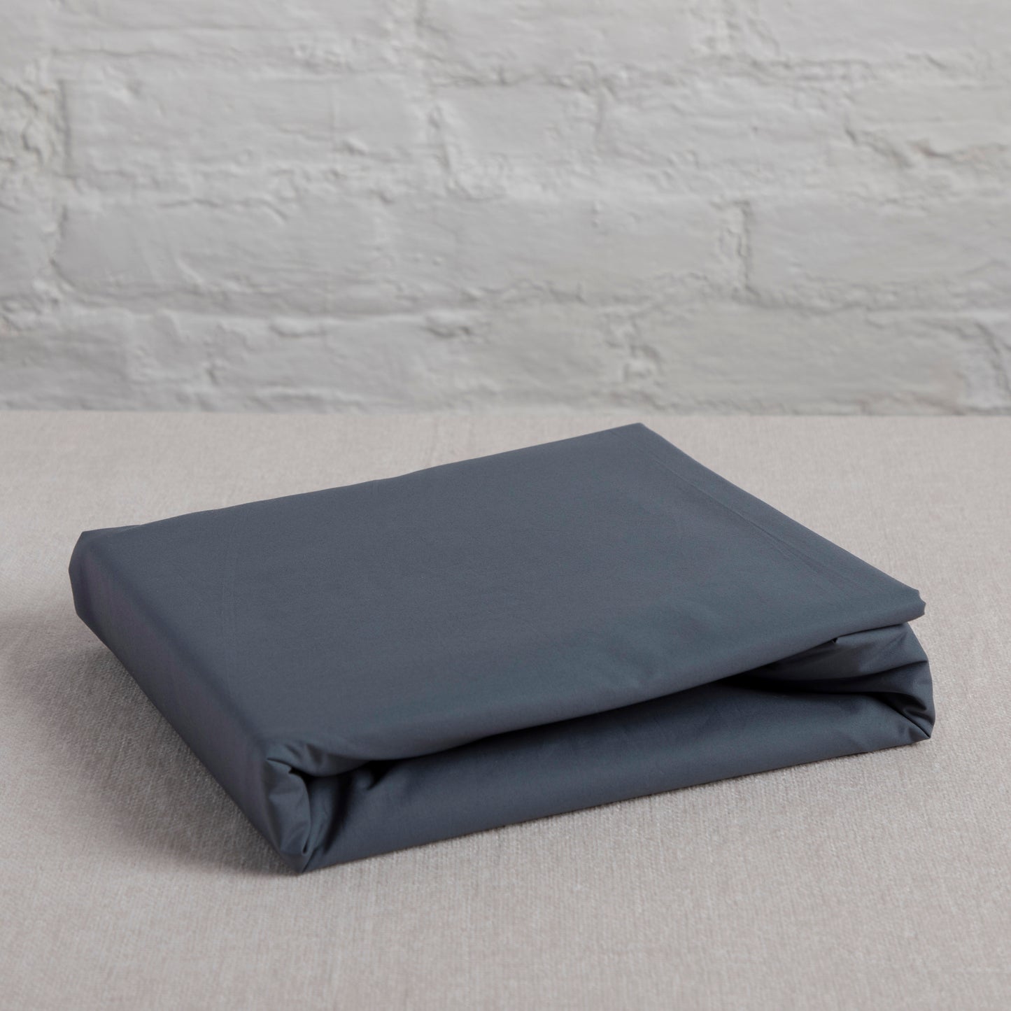 Folded sargasso blue organic pure cotton GOTS certified flat sheet on a light gray surface with a stone wall background