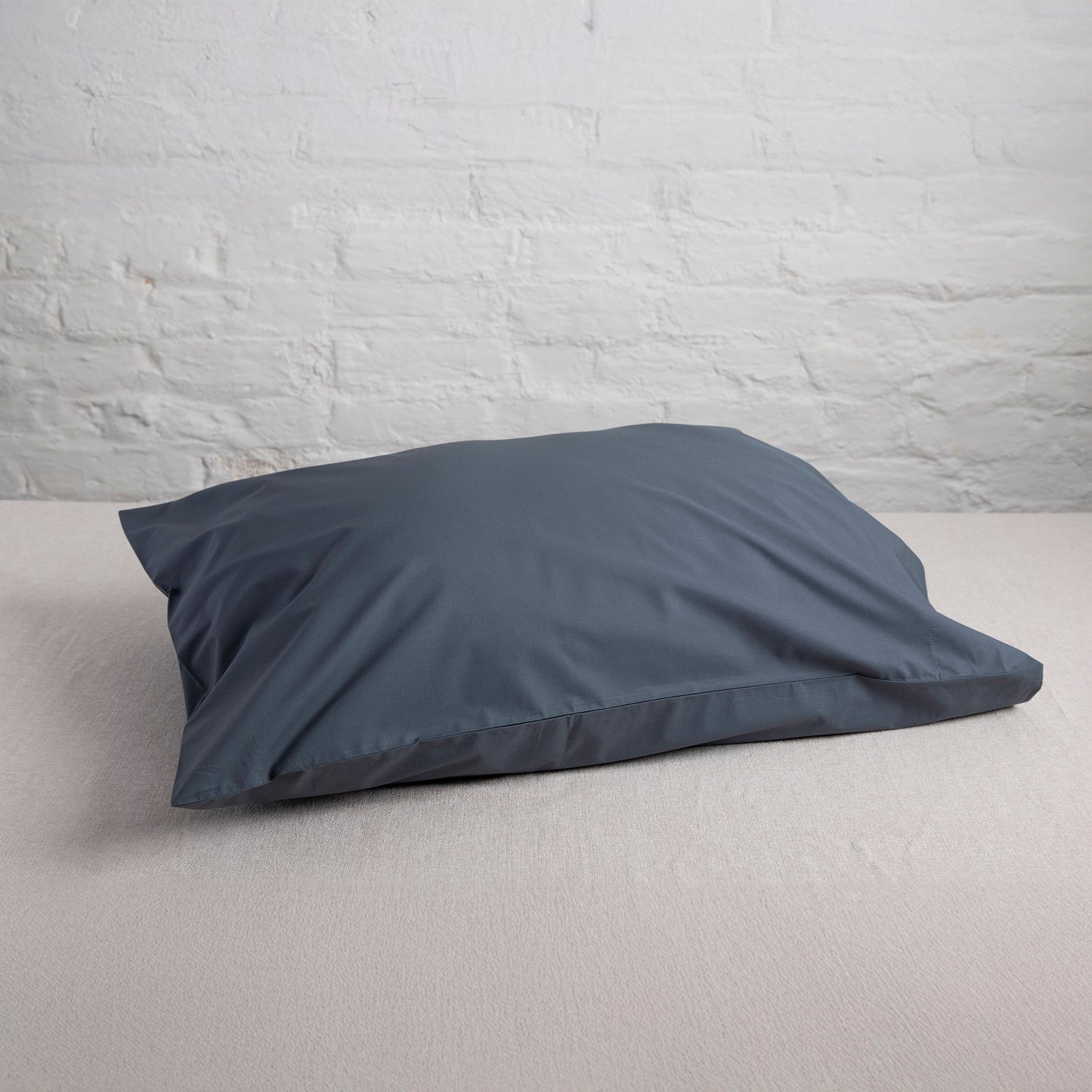Sargasso blue 100% organic cotton pillow on a light gray surface with a white brick wall background
