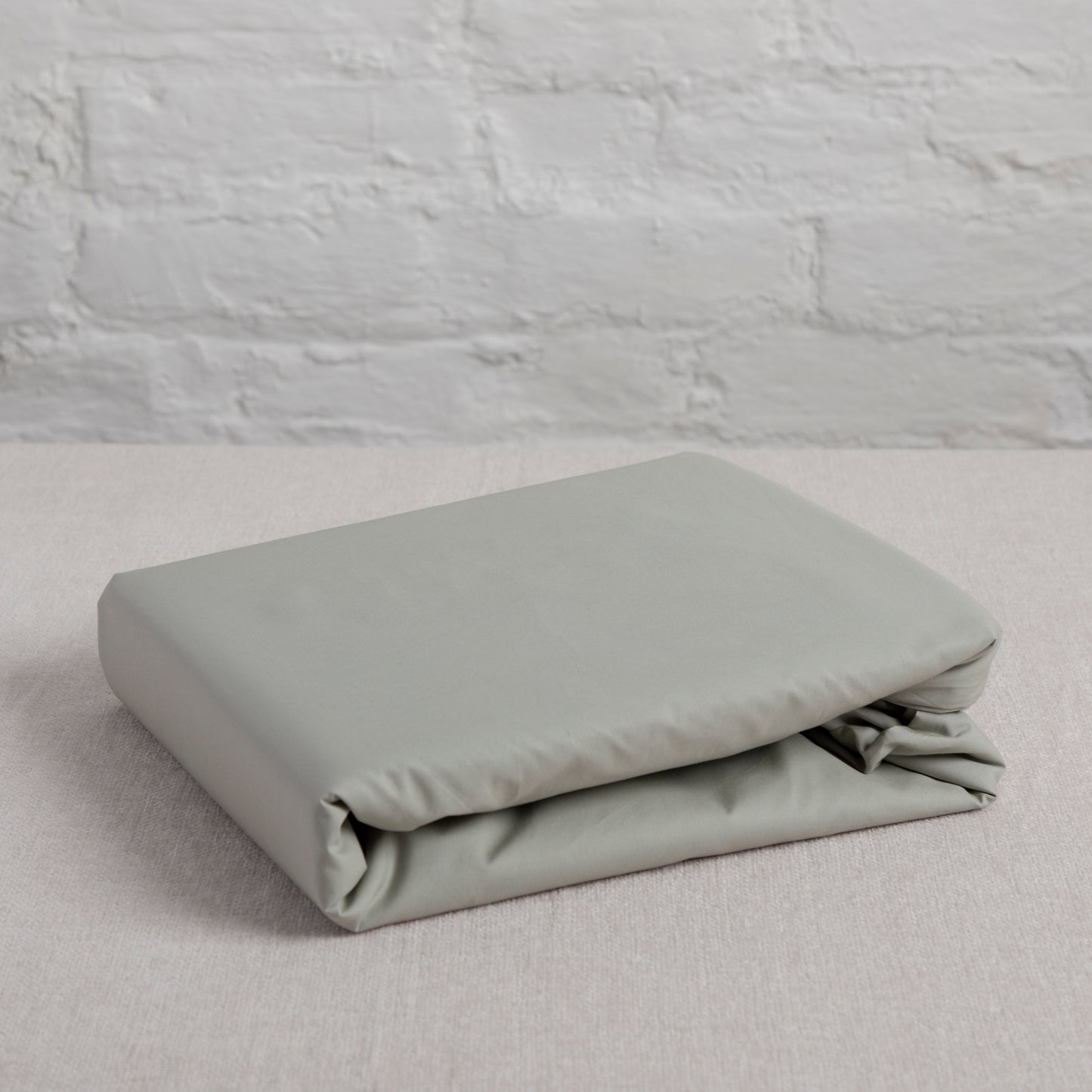 Folded Seagrass green 100% pure cotton flat bed sheet on a bed against a light gray brick wall.