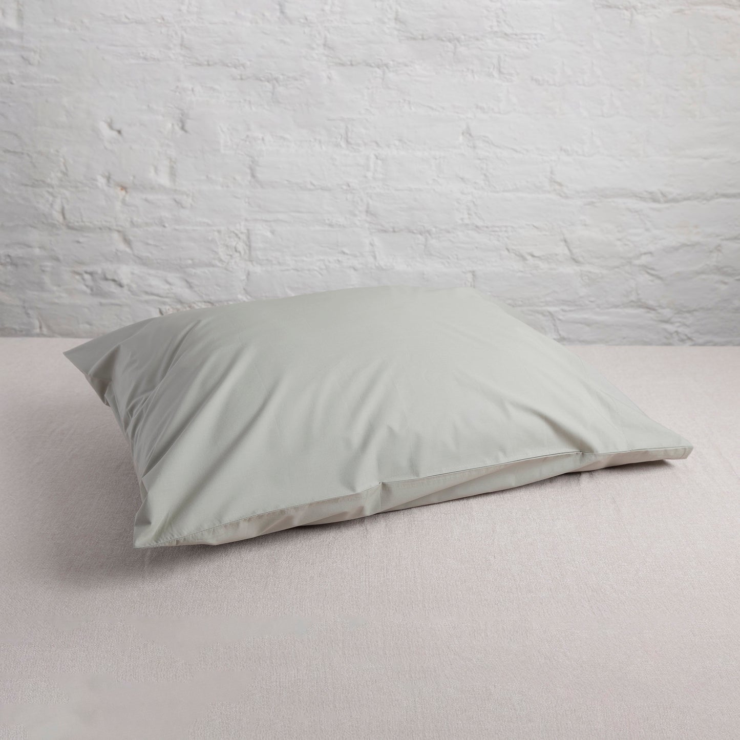 Seagrass green organic cotton pillow on a light grey bed with a white brick wall background