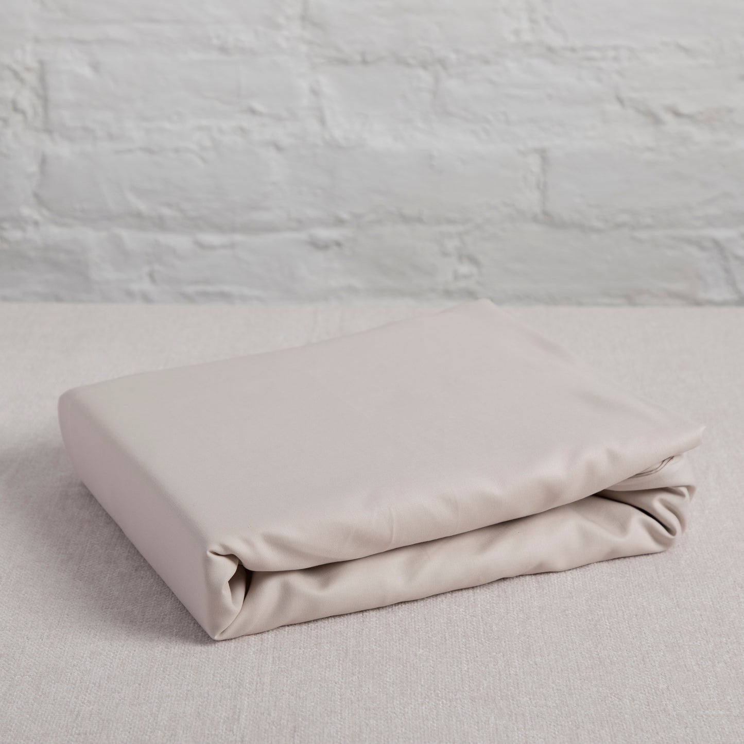 Folded beige driftwood natural cotton flat sheet on a light gray surface with a textured gray wall background – Linen Bay UK