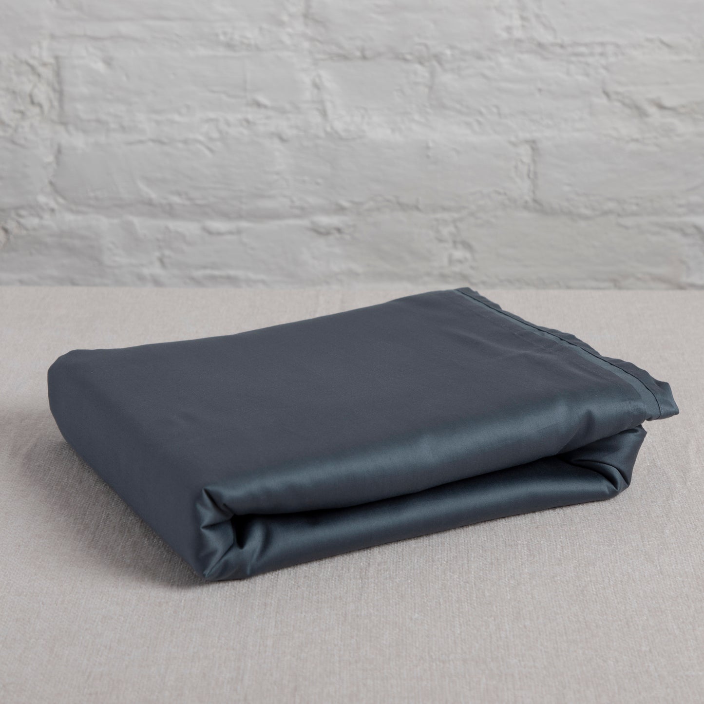 Folded sargasso blue cotton flat sheet on a light gray surface with a textured gray wall background – Linen Bay UK
