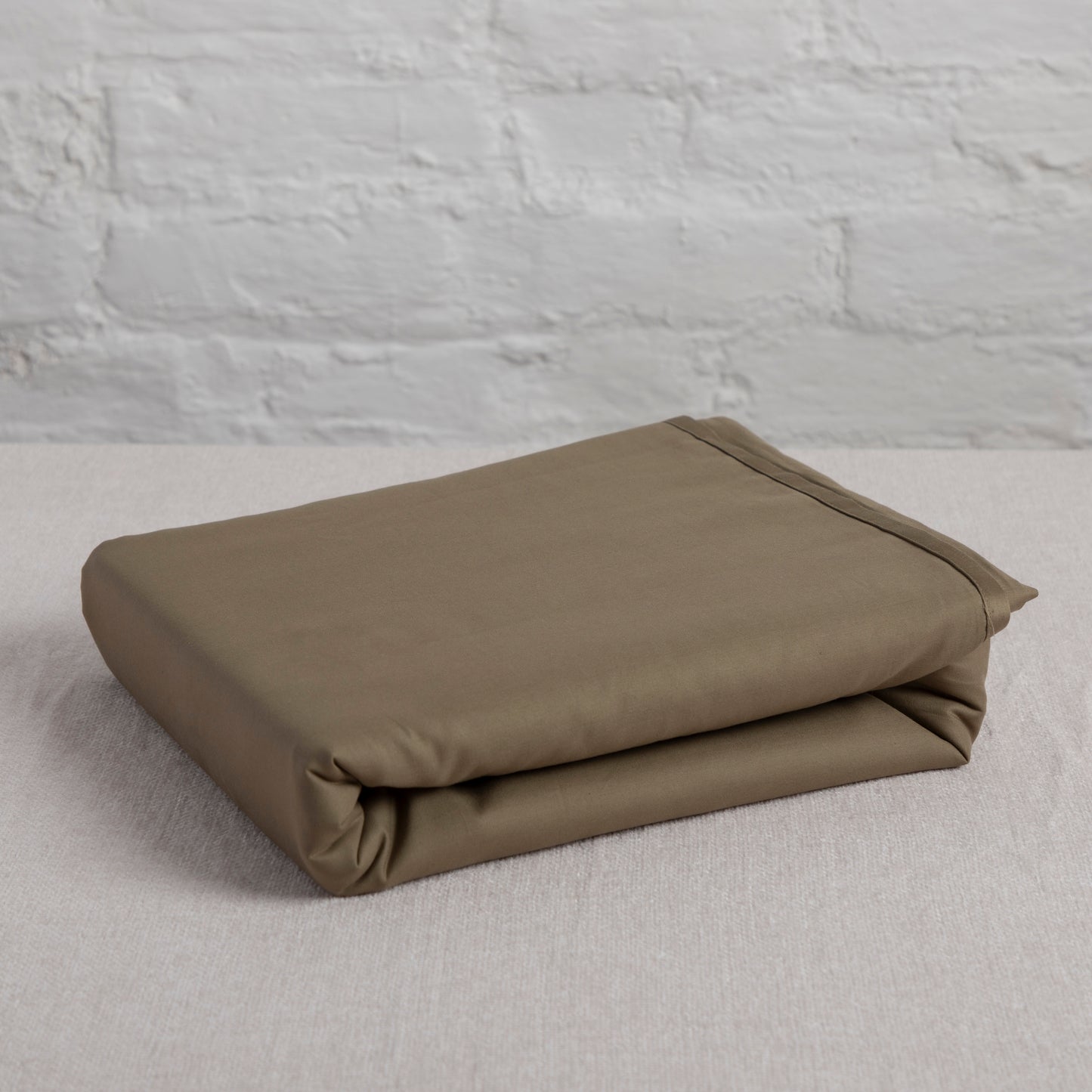 Folded seafern green cotton flat sheet on a light gray surface with a textured gray wall background – Linen Bay UK