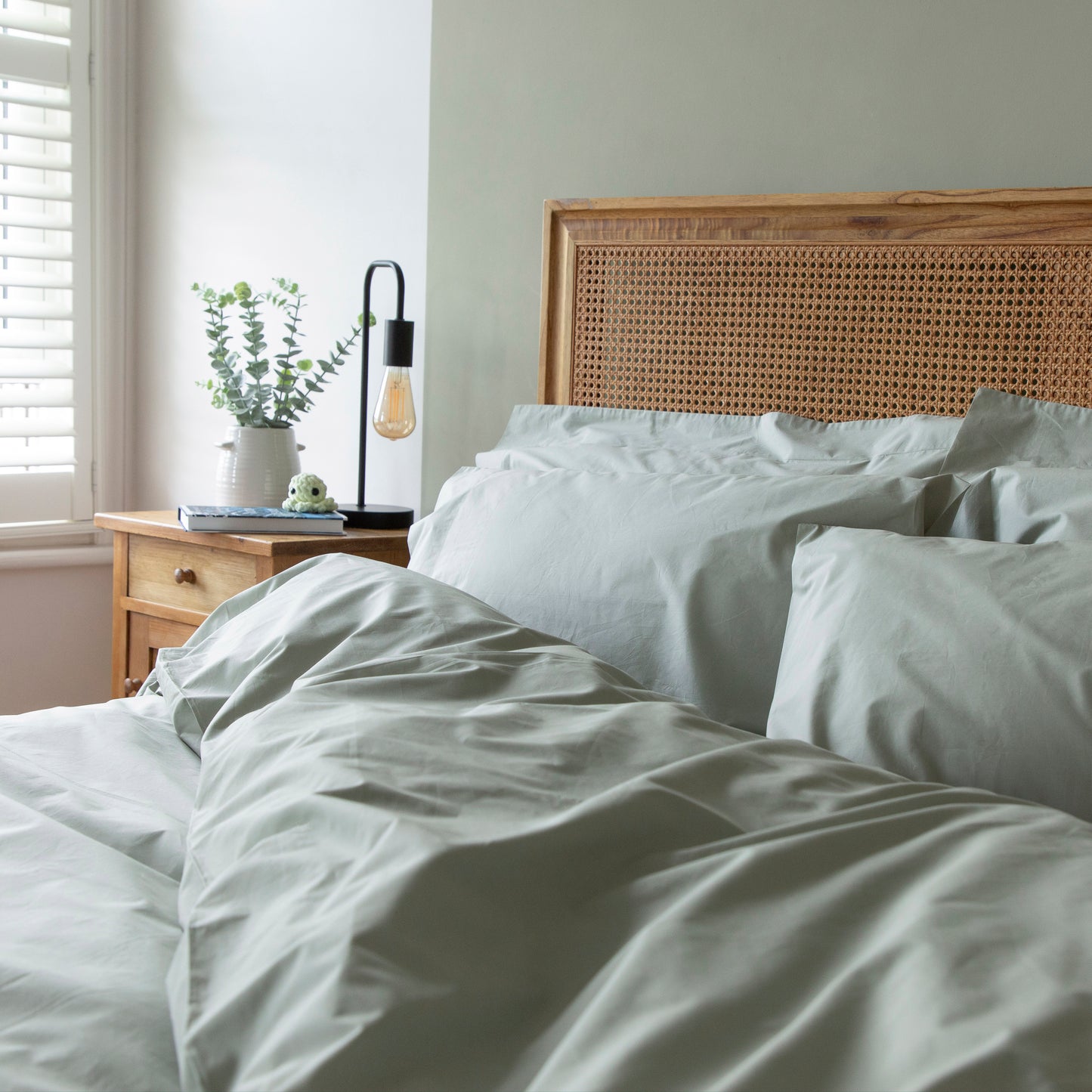 Bedroom with a bed featuring light-colored bedding and a wooden headboard, nightstand with lamp and plant – Linen Bay UK