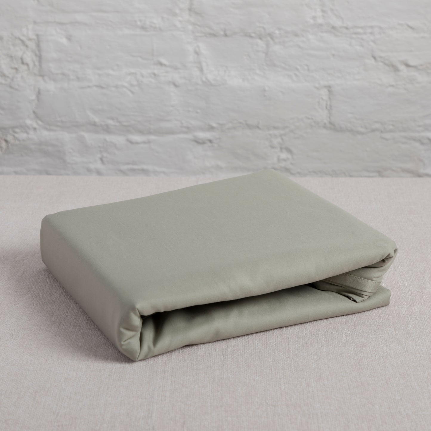 Folded beige seagrass green cotton flat sheet on a light gray surface with a textured gray wall background – Linen Bay UK