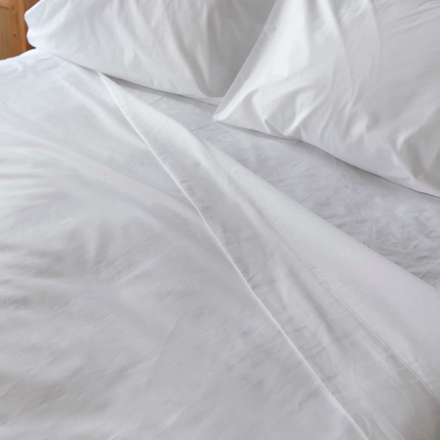 White bedding set with flat shet and pillows on a wooden bed frame.
